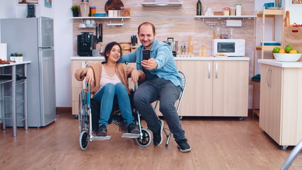 Positive woman in wheelchair and husband taking a selfie in kitchen using smartphone. Hopeful husband with handicapped disabled disability invalid paralysis handicap person next to him, helping her to reintegrate photo