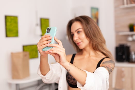 Portrait of beautiful woman looking at phone camera while taking selfie in home kitchen weraring sexy underwear. Seductive woman with tattoos using smartphone wearing temping lingerie in the morning. photo