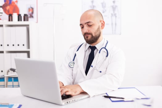 Doctor wearing stethoscope and white coat in hospital office using laptop to write patient treatment. Medical practitioner using notebook in clinic workplace , confident, expertise, medicine. photo