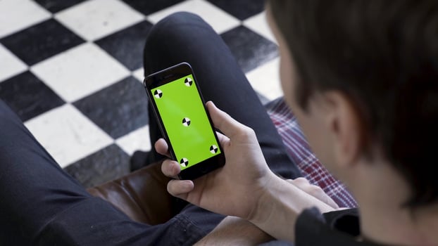 Over the shoulder view of a man using smart phone with green screen in the room with chessboard floor. Stock footage. Male holding his device with chromakey screen and looking at it. photo