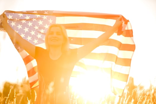 Beautiful young girl holding an American flag in the wind in a field of rye. Summer landscape against the blue sky. Horizontal orientation. photo