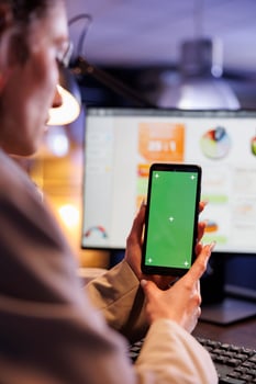 Entrepreneur looking at green screen mock up chroma key smartphone with isolated display, analyzing marketinf graphs report. Executive manager working late at night in startup office photo