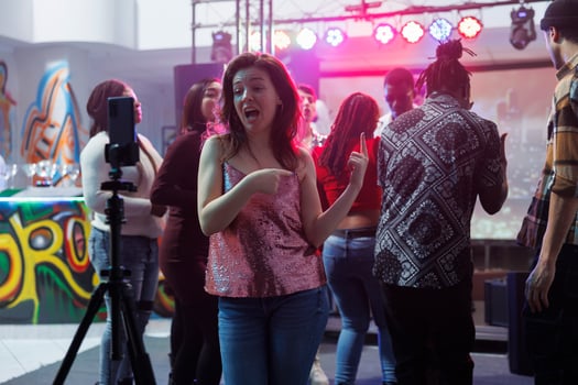 Carefree woman speaking on smartphone camera while recording video at nightclub party. Girl clubber broadcasting club social gathering online in social media using mobile phone on tripod photo
