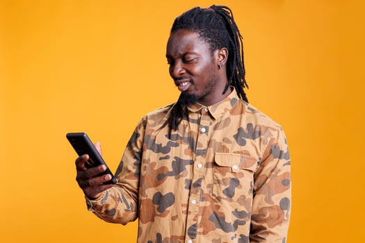 African american man reading job application on smartphone, scrolling through social media in studio over yellow background. Cheerful young adult searching information on mobile phone photo