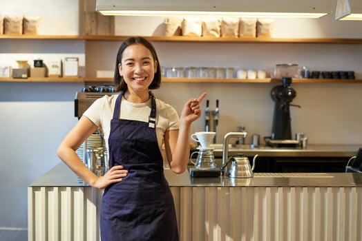 Portrait of asian girl in apron, standing in cafe near counter, barista pointing finger at copy space, empty banner coffee shop photo