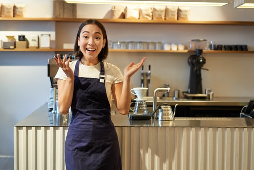 Enthusiastic young asian woman, barista in apron, looking amazed, clap hands and smiling, standing in coffee shop, working in cafe photo
