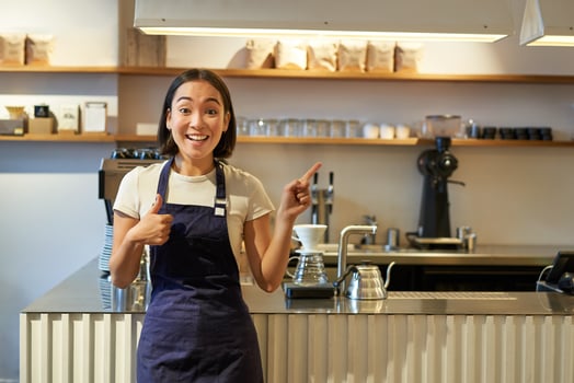Happy young asian barista, female staff worker in apron, showing thumbs up and pointing at copy space, demonstrating cafe advertisement photo