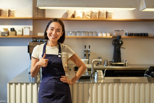 Smiling girl student working part-time in cafe, barista shows thumbs up, wears apron, stands near coffee shop counter photo