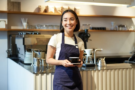 Portrait of asian smiling female barista, cafe waitress holding cup of coffee, serving clients, taking order to the tablet, standing near cafe counter photo