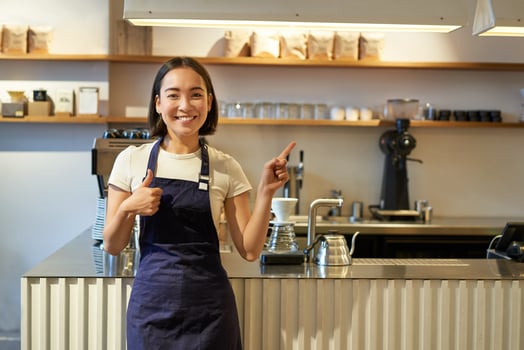 Happy young asian barista, female staff worker in apron, showing thumbs up and pointing at copy space, demonstrating cafe advertisement photo