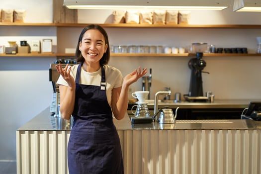 Enthusiastic young asian woman, barista in apron, looking amazed, clap hands and smiling, standing in coffee shop, working in cafe photo