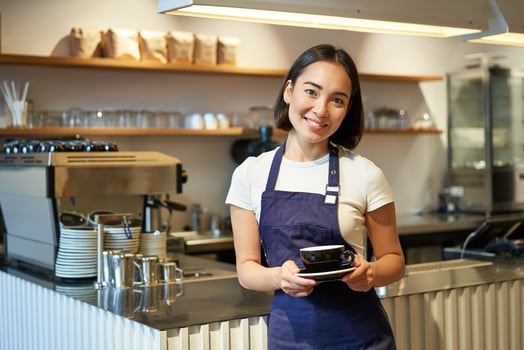 Portrait of asian smiling female barista, wearing cafe apron and holding cup of coffee, taking client order to the table, serving drinks photo