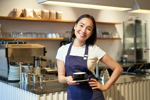 Portrait of smiling asian female barista, making coffee, holding cup of tea and taking it to cafe client, wearing apron, standing near counter photo