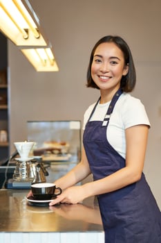 Vertical shot of smiling asian girl barista, wearing uniform, making coffee, standing near counter with cup, working in cafe photo