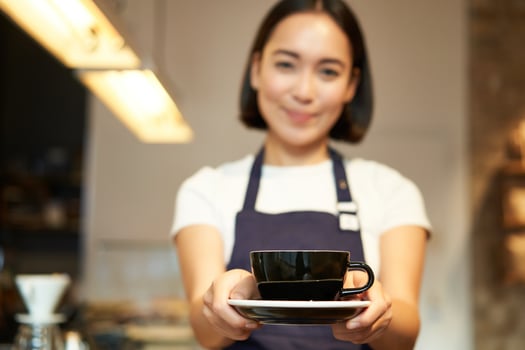 Smiling asian girl barista, standing in apron uniform, giving you cup of coffee, made a drink for client in cafe photo