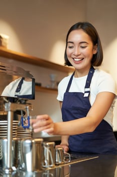 Portrait of cute barista girl working behind counter, making coffee, steaming milk for cappuccino, wearing cafe uniform blue apron photo