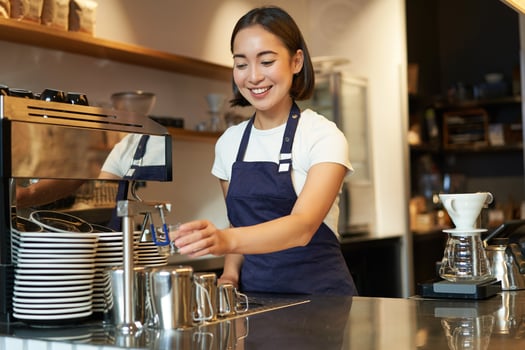 Smiling girl barista in cafe, preparing cappuccino in coffee machine, steaming milk, wearing uniform apron photo