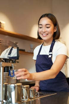 Portrait of cute barista girl working behind counter, making coffee, steaming milk for cappuccino, wearing cafe uniform blue apron photo