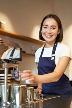 Vertical shot of young professional, smiling girl barista working in cafe, steaming milk for cappuccino drink, preparing order with coffee machine photo