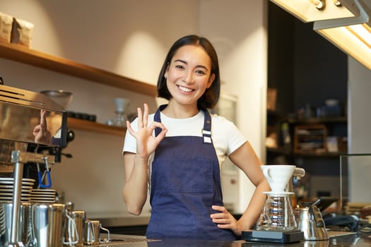 Barista at the counter shows okay sign. Smiling asian girl works in cafe, wears blue apron, recommends her coffee shop photo