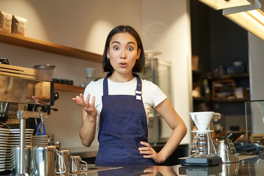 Portrait of girl barista in apron, looks surprised, points with hand and stares amazed at camera, works behind counter and brews coffee photo