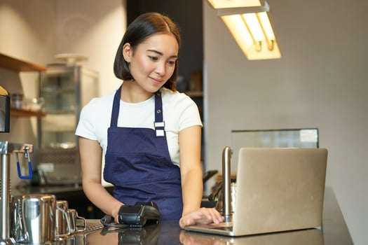 Smiling asian girl barista, working in cafe, serving client, looking at laptop while using card reader to receive money for order, selling coffee photo