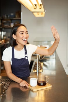 Smiling asian girl barista calling client, giving out order in cafe, prepared batch brew filter coffee, wearing apron behind counter photo