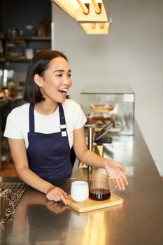 Smiling asian girl barista calling client, giving out order in cafe, prepared batch brew filter coffee, wearing apron behind counter photo