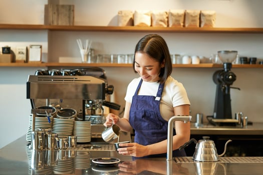 Happy smiling cafe owner, girl barista in apron, making cappuccino, latte art with steamed milk, standing behind counter photo