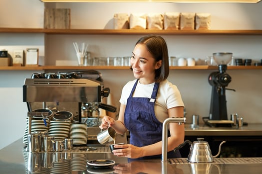 Happy smiling cafe owner, girl barista in apron, making cappuccino, latte art with steamed milk, standing behind counter photo