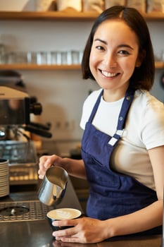Vertical shot of friendly barista girl, smiling at you while preparing coffee, pouring milk and making cappuccino photo