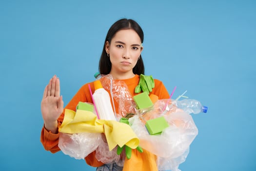 Serious eco activist shows stop sign and plastic waste, urges to stop throwing recycling materials into general garbage bins, blue background photo