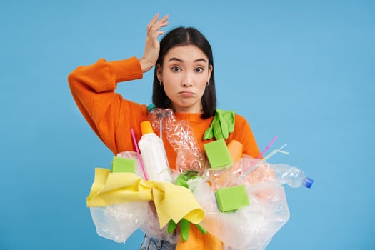 Confused Korean woman tries to go green, recycle plastic household items, holding bottles and gloves, does recycling, blue background photo