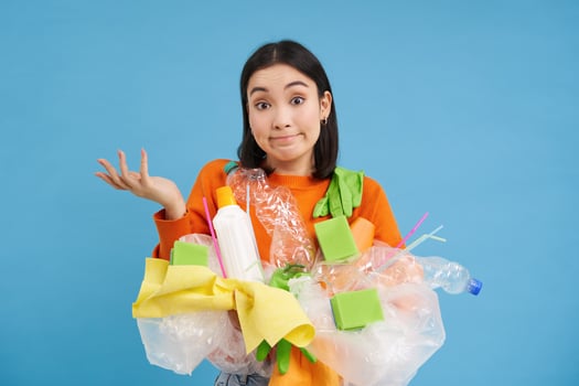 Korean girl with plastic garbage, shrugs shoulders, doesnt know how to recycle, sorting her waste for recycling station, blue background photo