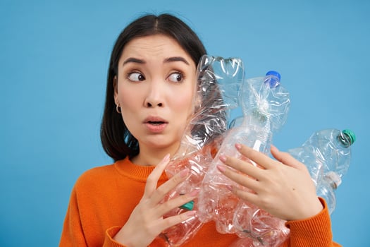Portrait of asian girl with scared face, holding piles of plastic bottles for recycling and stares to the right with shocked emotion, blue backgrond photo