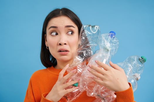 Portrait of asian girl with scared face, holding piles of plastic bottles for recycling and stares to the right with shocked emotion, blue backgrond photo