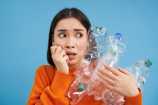 Portrait of asian girl with scared face, holding piles of plastic bottles for recycling and stares to the right with shocked emotion, blue backgrond photo