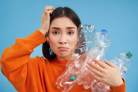 Sad asian woman holding plastic bottles, looking troubled and upset, sorting waste for recycling, standing over blue background photo