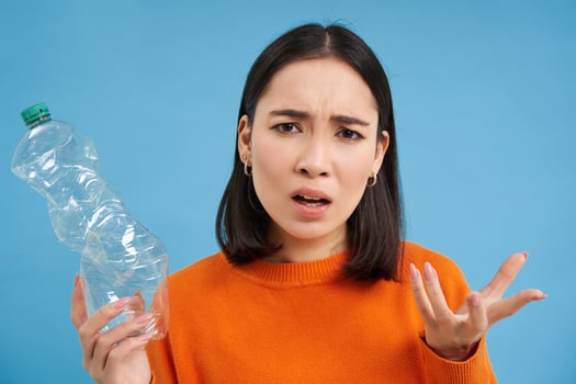 Portrait of confused woman, holding and showing plastic bottle, clueless why people are not recycling, blue background photo