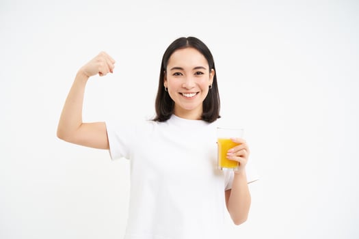 Healthy lifestyle. Smiling young asian woman, shows strong muscles, biceps on hand and drinks orange juice, white background photo