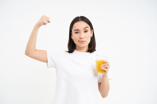 Healthy lifestyle. Smiling young asian woman, shows strong muscles, biceps on hand and drinks orange juice, white background photo