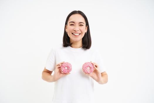 Funny korean woman posing with two delicious glazed doughnuts on chest, laughing and smiling, eats junk food, tasty dessert photo