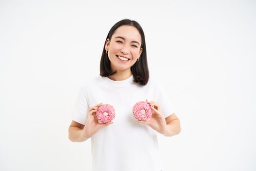 Smiling happy asian woman laughing, holding two glazed doughnuts over chest, standing over white background photo