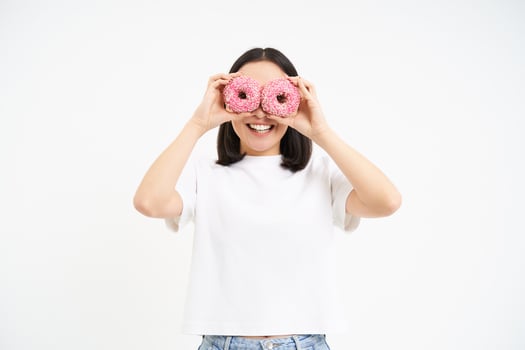 Portrait of happy korean woman, makes glasses from two tasty glazed doughnuts, smiling and having fun, white background photo