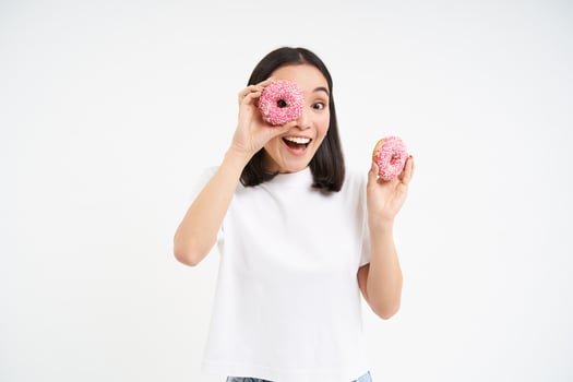 Portrait of happy korean woman, makes glasses from two tasty glazed doughnuts, smiling and having fun, white background photo