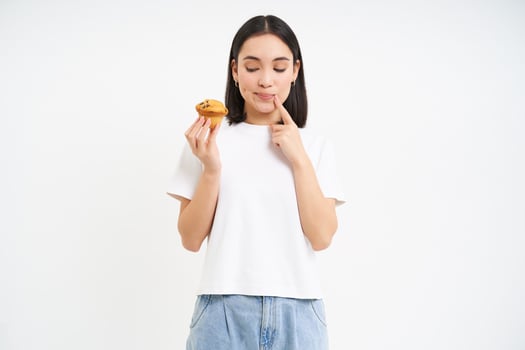 Bakery and sweets. Happy asian woman looking at tasty cupcake, eating pastry, white background photo