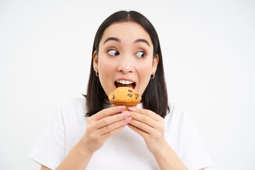 Close up of happy korean woman eating cupcake, delicious dessert, smiling from pleasure, white background photo