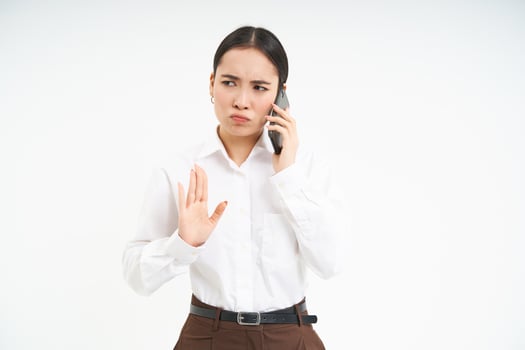 Portrait of serious korean woman, business professional shows stop sign, talks on mobile phone with disapproval, frowns upset, stands over white background photo