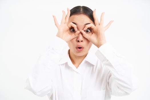 Business people. Happy asian girl looks through finger glasses, stares with surprised face, stands isolated on white background photo