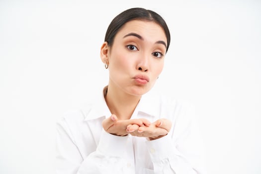 Close up portrait of cute office lady sends air kiss on palms, puckers lips and looks tender at camera, stands over white background photo